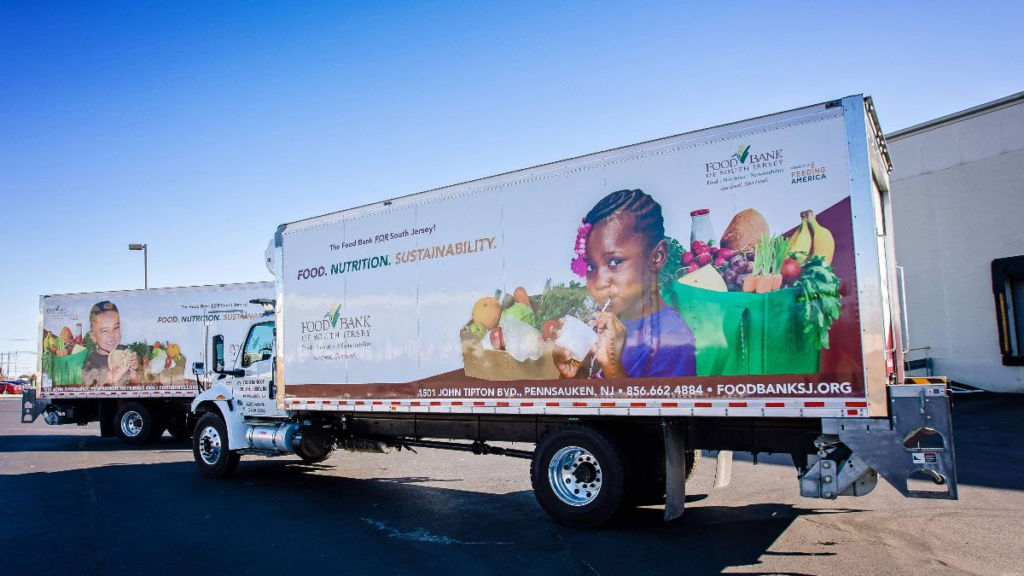 Photo of a tractor trailer featuring a young Black girl drinking from a carton. Behind her are fruits and vegetables inside of a brown box and a green reusable grocery bag. The words "The Food Bank FOR South Jersey" and "Food. Nutrition. Sustainability" are also seen, as well as the Food Bank of South Jersey logo. Additional contact information, such as the physical address, phone number and website, is also featured