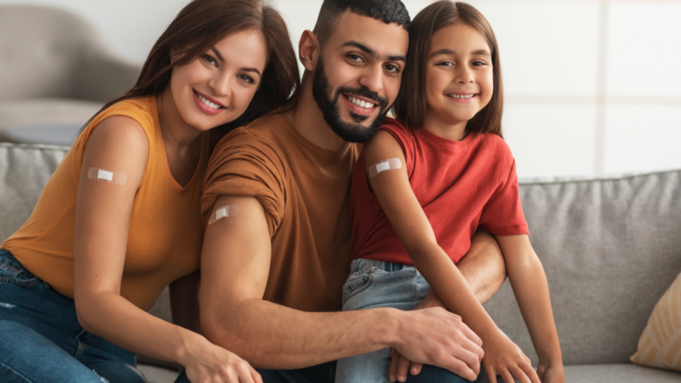 COVID vaccine family Middle Eastern family - mother, father, daughter - sitting on a tan couch, smiling at the camera, showing their right arms with adhesive bandage