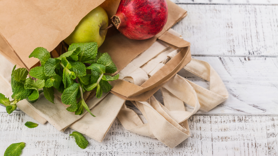 Green Environment Reusable Bag Brown bag with some leafy greens, a green apple, and a red pomegranate on top of tan reusable bags. The background is a white-washed wood.
